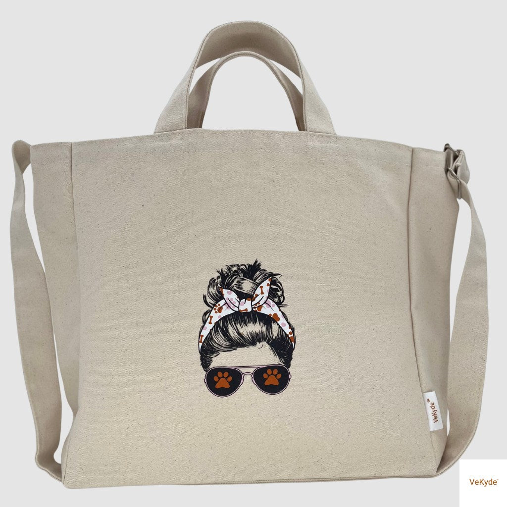 Natural canvas tote bag featuring a printed design of a woman with a messy bun, dog-themed headband, and sunglasses with paw prints. Eco-friendly pet lover gift by VeKyde